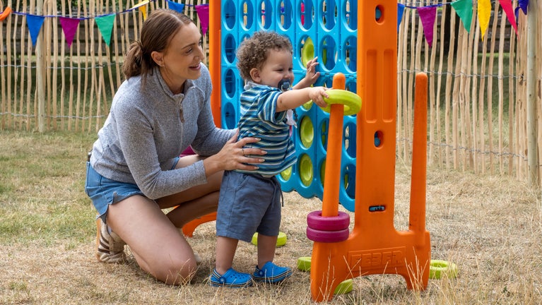 A child standing next to a crouching adult playing with a giant connect 4 game in a grass activities area at Summer of Play